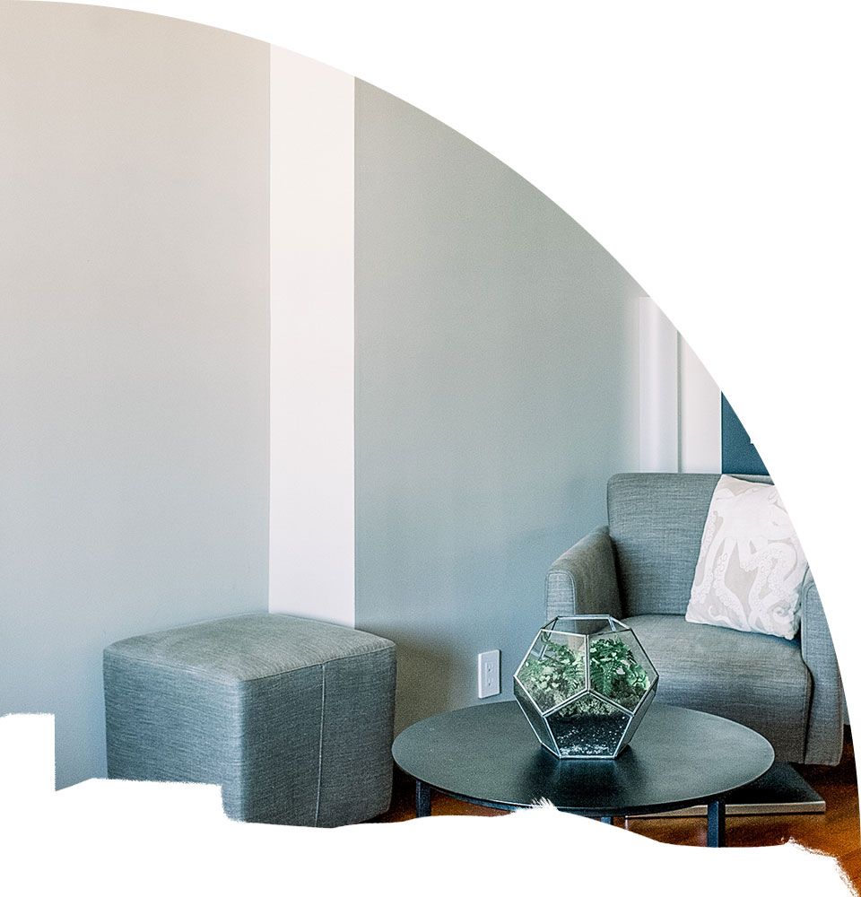 Fotografía de parte de un salón con un sillón, una mesa y una pared de fondo acabada con pintura gris claro